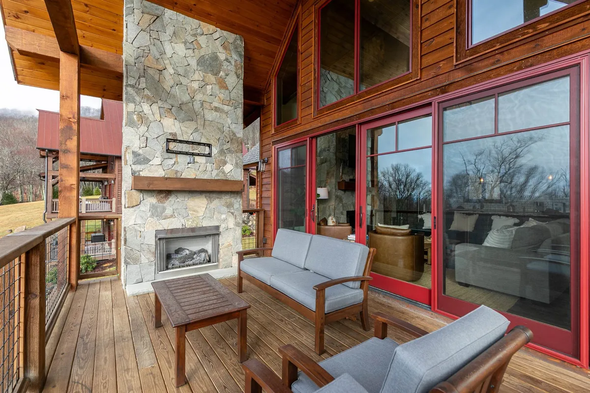 A cozy exterior patio scene featuring a stone fireplace, comfortable wooden patio furniture, and expansive glass windows offering views of the natural landscape. The scene suggests relaxing after a day’s adventure, perhaps enjoying the sunset or gathering around the fire on a cool night.