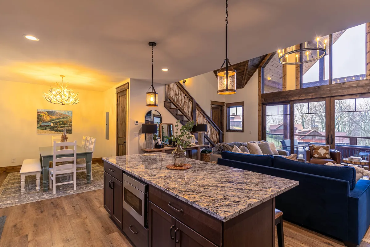 This image captures the heart of the social space: a granite kitchen island at the center, a dining area with white chairs, a cozy blue couch seating area, and large windows filling the room with natural light. The open layout allows seamless flow between kitchen, dining, and living areas, while mountain-inspired decor and plush furnishings create a welcoming atmosphere.
