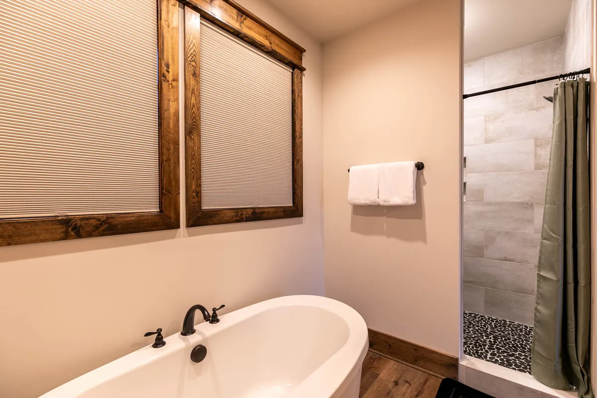 A serene bathroom scene with a freestanding bathtub, glass shower area, and wooden window frames. Natural light streams in, highlighting fresh towels and a sense of spa-like tranquility. The clean, well-organized vanity hints at a starter set of toiletries for guest convenience.