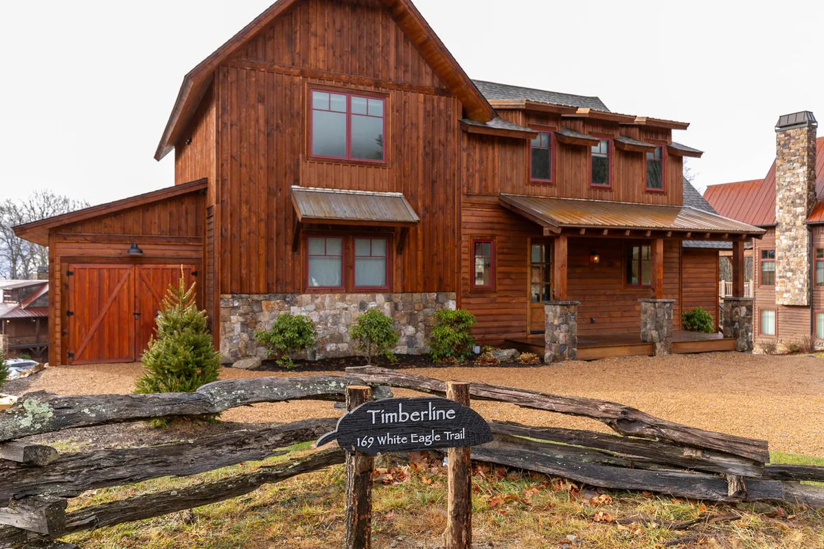 The image showcases the exterior of Timberline at Eagles Nest, featuring a striking wooden facade with elegant stone accents and expansive windows that frame the sunset. The welcoming porch hints at cozy evenings and unforgettable mountain views, while tasteful signage affirms a sense of arrival.