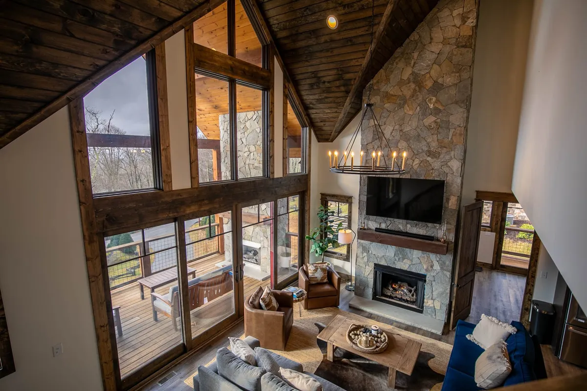 A warm, inviting living area featuring a large stone fireplace, comfortable seating, floor-to-ceiling windows drawing in the natural landscape, and rustic wooden beams overhead. The scene suggests cozy evenings relaxing by the fire or enjoying the sunrise through expansive glass.