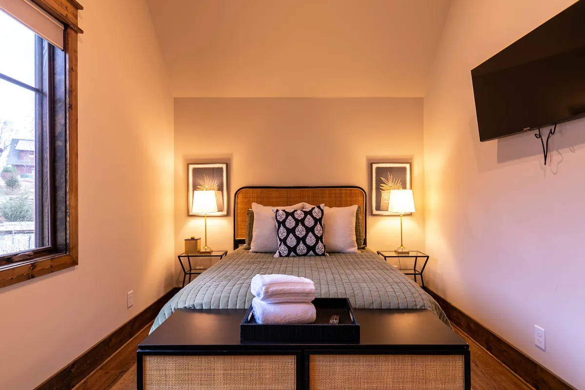 A cozy bedroom featuring a comfortable bed with decorative pillows, nightstands, soft lamps, and wall art. The room is bathed in natural light, evoking a peaceful and restful setting suitable for couples or families.